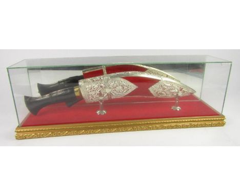 A Gurkha ceremonial kukri, the knives with horn handles, scabbard of red felt with white metal overlay cast with peacocks, fl