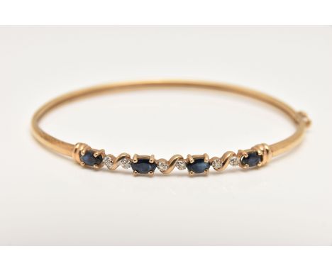 A YELLOW METAL GEM SET BANGLE, set with four oval cut deep blue sapphires interspaced with illusion set single cut diamonds, 