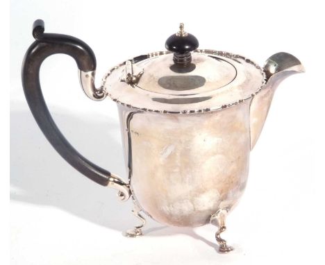 George V silver teapot of circular form having a hinged lid with an ebonised urn finial and scroll handle, the border applied