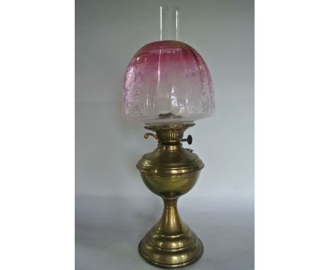 A brass oil lamp supporting a clear glass chimney and a cranberry tinted shade of domed form with etched foliate decoration, 