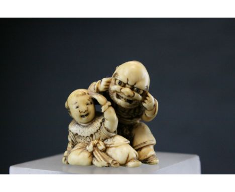 An ivory netsuke of father and son playing, signed, the kneeling father wearing a noh mask, while the boy shields his eyes, H