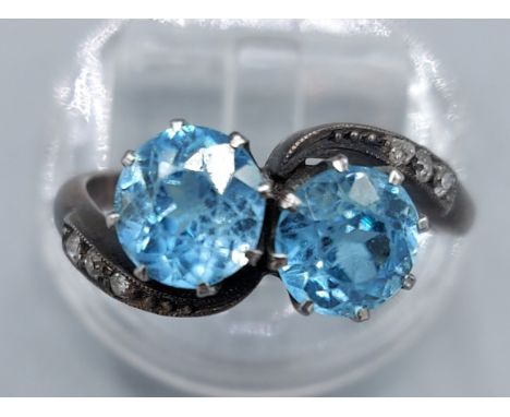 A white metal blue stone and diamond ring set with two blue stone within a crossover setting with diamond shoulders, ring siz