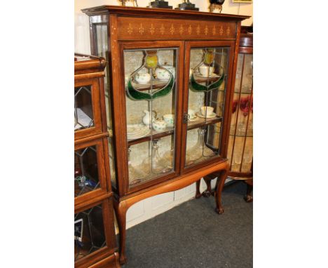 An Art Nouveau inlaid mahogany display cabinet with two shelves enclosed by two lead glazed panel doors decorated with styliz