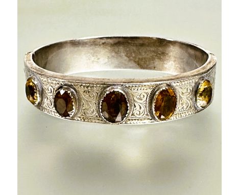 A late Victorian white metal engraved stiff hinged hollow bangle with five oval colette set graduated citrines and and cairng