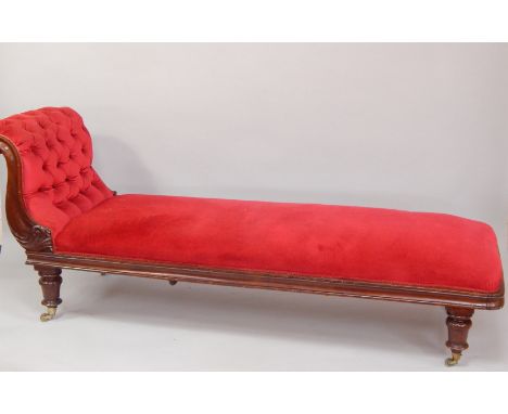 A Victorian mahogany chaise longue, upholstered in red button back draylon, with a leaf carved scrolling arm, raised on turne