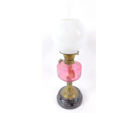 An early 20thC brass oil lamp, with a cranberry glass reservoir raised on a black pottery socle, with chimney and white globu
