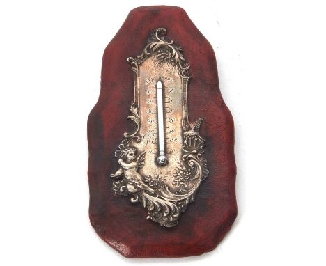 A late Victorian silver mounted thermometer on a red leather frame, embossed and chased with cherubs, scrolls and foliage, 16