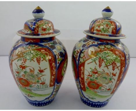 A PAIR OF JAPANESE IMARI VASES with covers, painted in iron red, blue, green and gilded, having floral panel decoration, 26cm
