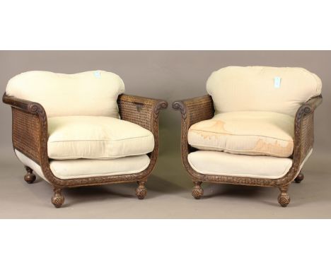 A pair of George V mahogany framed bergère armchairs, the frames carved with leaf sprays, each with cream cotton loose cushio
