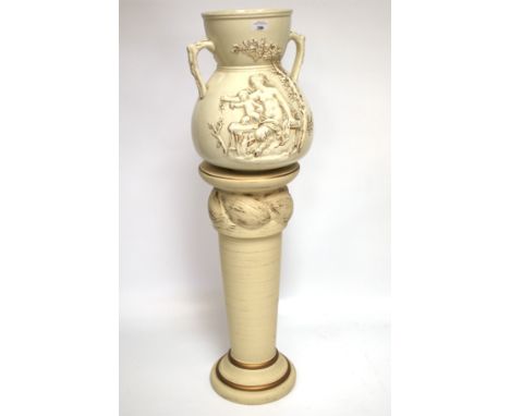 A large cream glazed stoneware pot with twin handles on a modern ceramic pedestal base. The pot moulded with woman and child 