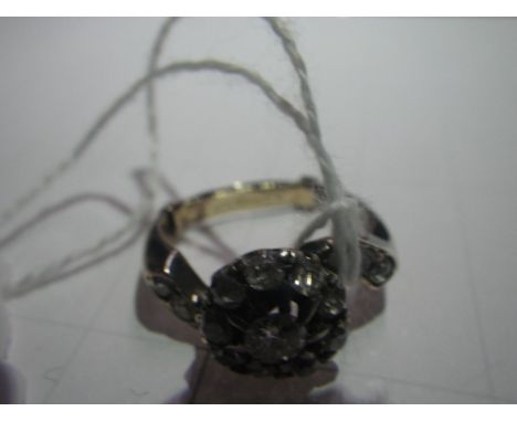 A DIAMOND CLUSTER RING, in the form of a flower with twisted shoulders set with diamonds, set on an unmarked white metal shan