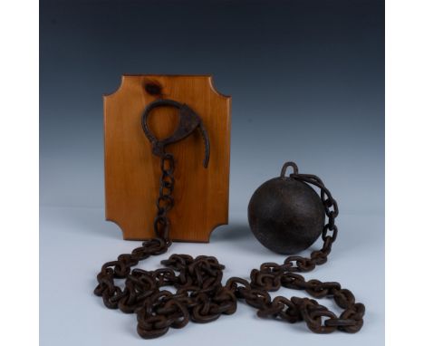 Presents a very heavy iron ball with chain shackle, marked 25 pounds. The shackle is mounted on a polished wooden plaque, Pla
