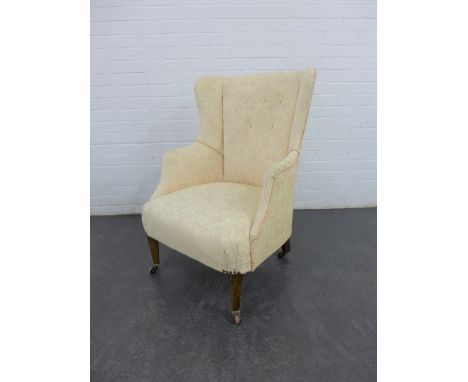 An upholstered wing back armchair with buttonback, on tapering mahogany legs with brass caps and ceramic castors, 93 x 70 x 6