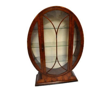 An Art Deco walnut display cabinet of oval form, the single glazed door enclosing two fixed shelves, raised on plinth base, h