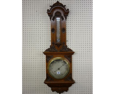 A CARVED OAK ANEROID WALL BAROMETER, elaborately framed with silvered dial having a brass bezel, a mercury thermometer above 