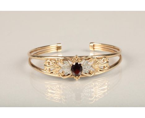 9ct gold and garnet set bangle, the central oval cut garnet flanked by illusion set diamonds on a baroque style openwork fram