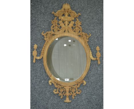 An oval wall mirror, the pediment pierced and carved with putti holding aloft an urn amid flora and swags.