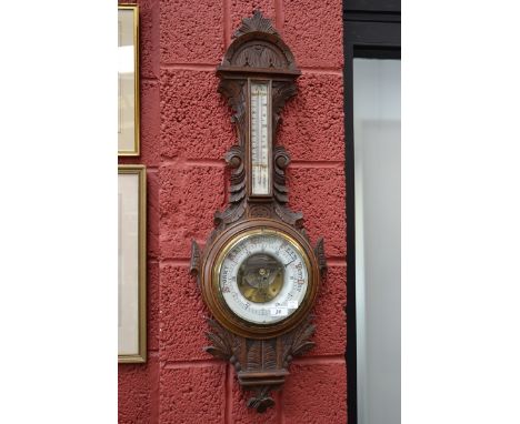 An Edwardian carved oak aneroid barometer, exposed workings set in a ceramic dial, mercury thermometer,  
