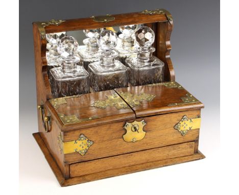 A late Victorian oak and brass mounted three bottle tantalus, applied with gothic brass mounts and side swing handles, the mi
