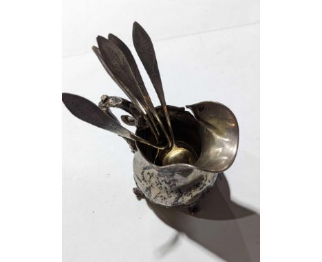 A short silver or silver-plated jug alongside five plated spoons. The jug smooth and round, and upon four legs, with handle r