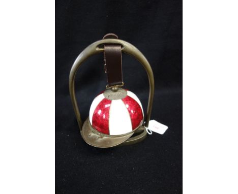 AN ENAMEL AND BRASS TABLE BELL in the form of a jockey's cap, supported by a brass stirrup shaped stand, 21cm high