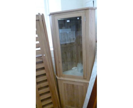 A modern light oak freestanding corner display cabinet with a single glazed door, enclosing two glass shelves above a panelle