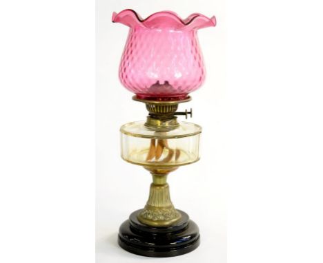A VICTORIAN BRASS OIL LAMP WITH FACETED GLASS FOUNT AND A DIMPLED CRANBERRY GLASS SHADE, ON BLACK GLAZED POTTERY FOOT, 45CM H