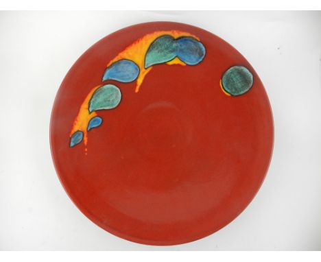 A Poole pottery Odyssey charger, having splashes of green and orange to one side upon a red ground. D.41cm