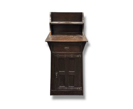An Arts and Crafts style cupboard of tapered form with raised shelf section over a single drawer and panelled cupboard with i