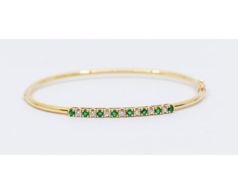 A diamond and emerald 14ct gold hinged bangle, comprising a row of fifteen alternate round mixed cut emeralds and brilliant c