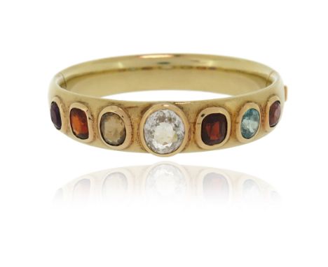 A gem-set gold bangle, the plain gold bangle is set with oval and cushion-shaped gemstones including garnets, zircons, a topa