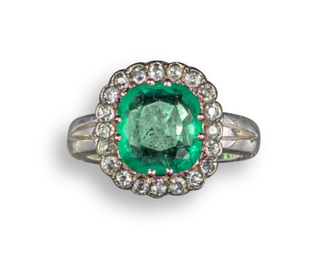 An emerald and diamond cluster ring, the cushion-shaped emerald is set within a surround of small circular-cut diamonds in pl