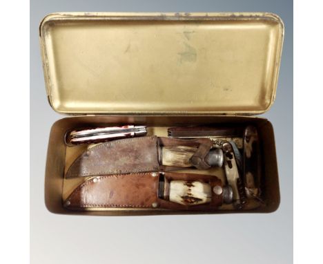A tin containing a collection of pocket knives and hunting knives in sheathes.