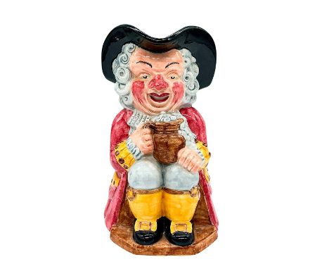 A hand painted figural ceramic jug depicting a seated man in a long pink coat with yellow breeches, and a black tricornered h