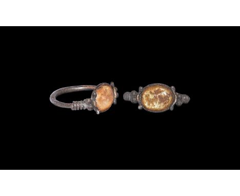 3rd-4th century AD. A Roman silver ring with coils and pellets to shoulders; the amber-tinted oval glass intaglio engraved wi