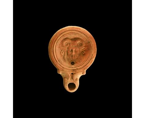 1st century AD. A ceramic oil lamp with rounded nozzle and volutes to the side; dish decorated with facing bust of a satyr wi