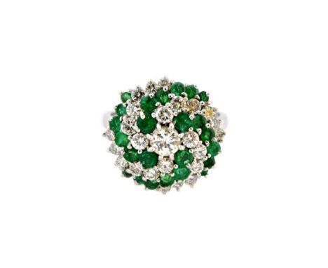 A white gold, emerald and diamond circular cluster ring, centred by a round brilliant cut diamond with spiralling settings of