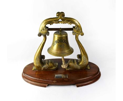 A brass ship's bell inscribed 'Demodocus' and '1962', raised on scrolling fish supports to oval wooden plinth, with applied p