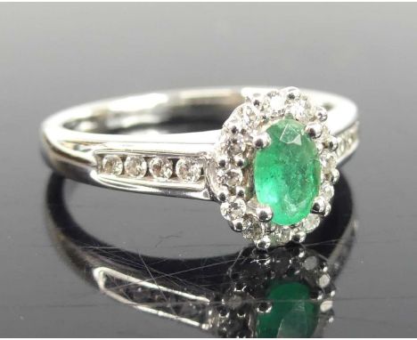 An 18ct white gold, emerald and diamond oval cluster ring, featuring a centre oval emerald within a border of twelve round br