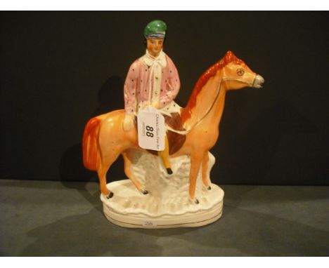 A Victorian Staffordshire mantle ornament, fashioned as a jockey wearing green cap and pink silks, seated upon a racehorse wi