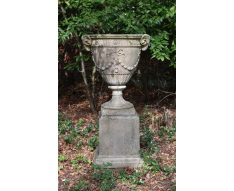 A CAST STONE URN IN THE NEOCLASSICAL STYLE, 20TH CENTURY  The urn with twin ram head handles with ribbons and swags, plinth b