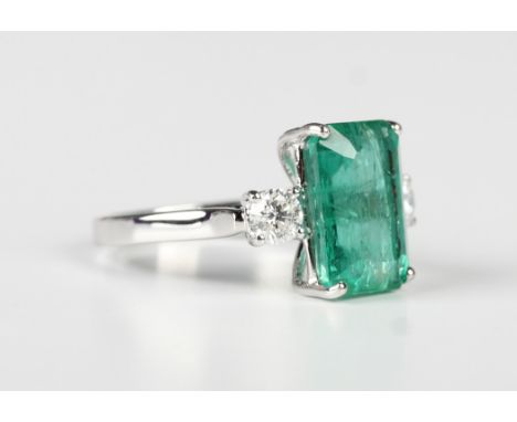 A white gold, emerald and diamond three stone ring, claw set with the cut cornered rectangular step cut emerald between two c