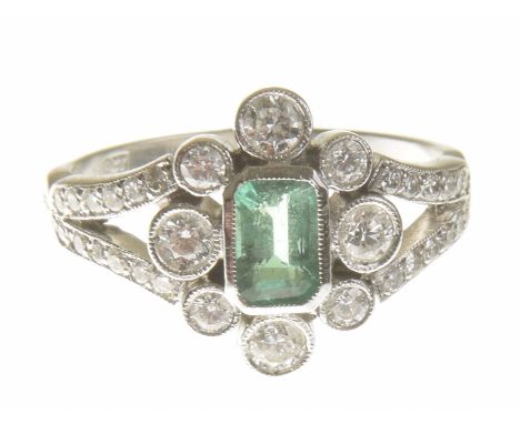 An emerald and diamond cluster ringThe rectangular shape emerald, within a vari size brilliant cut diamond collet surround, a