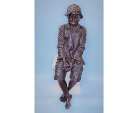 Frederich Goldscheider, a painted terracotta, life size, seated young boy, wearing a cap, hands clasped together and feet cro