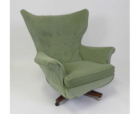 A 1960s upholstered winged swivel armchair, with cushion seat, raised on rosewood X-base to roller castors, w.90cm