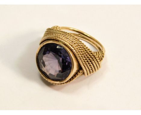 DRESS RING. A dress ring set a large synthetic colour-change sapphire within an unmarked yellow metal mount with braided deco