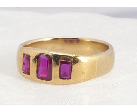 A three stone yellow metal ruby ring,  the three step cut rubies in sunken settings, on a wide setting and tapering shank, ri