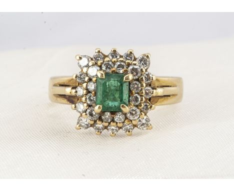 An 18ct gold emerald and diamond cluster ring, the claw set emerald surrounded by two bands of brilliant cut diamonds on a pl