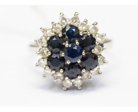 A white metal sapphire and diamond cluster ring, set with seven blue sapphires to the centre with smaller claw set diamond su