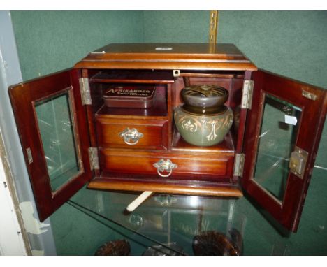 AN EDWARDIAN SMOKERS CABINET WITH A TWO DOOR GLAZED FRONT AND TWO DRAWERS, complete with a pipe rack and ceramic pottery jar